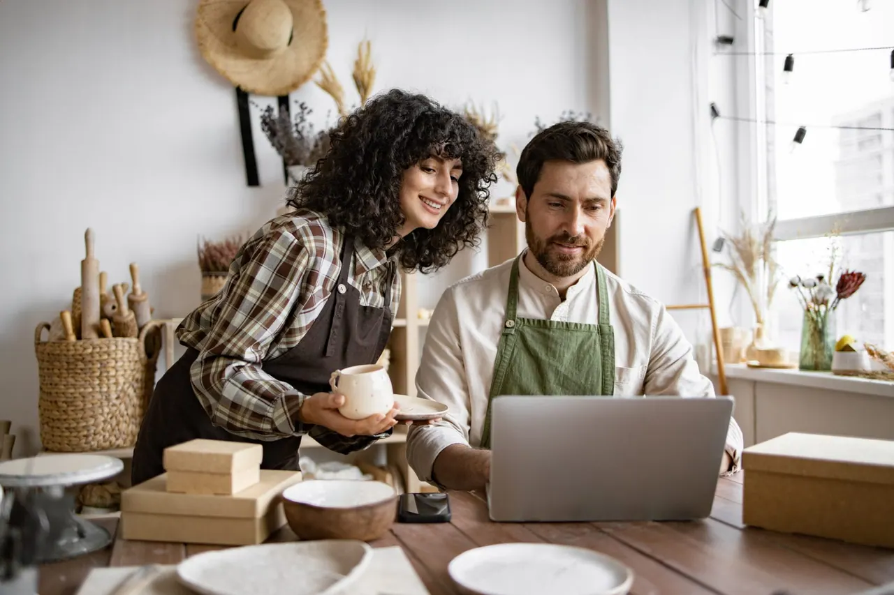 Two small business owners in aprons reviewing their website on a laptop in a pottery studio